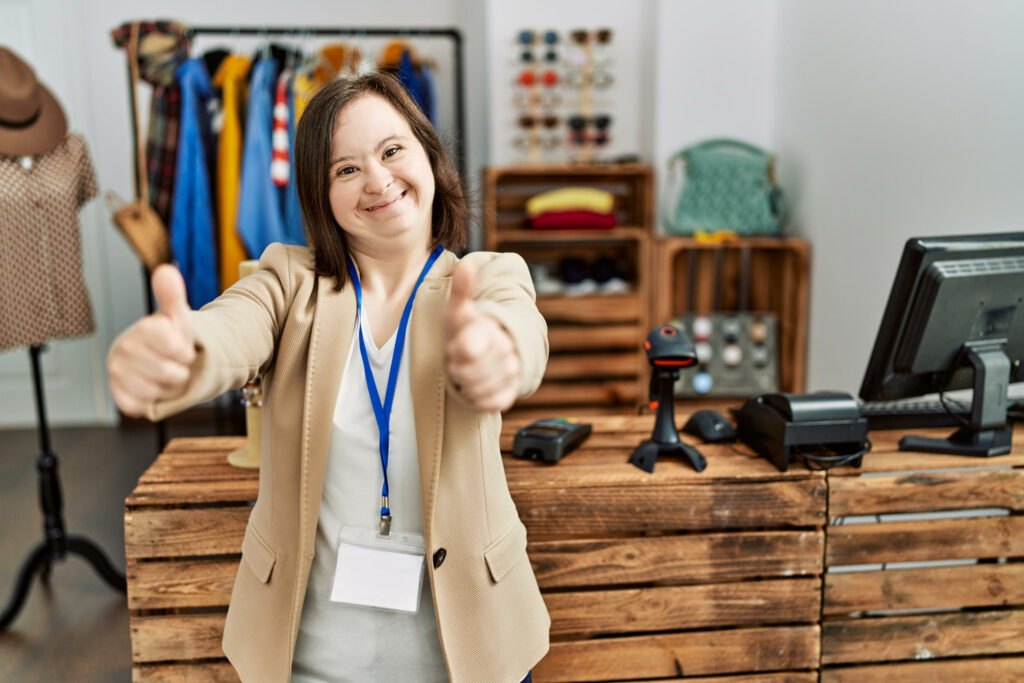 Young woman with developmental disabilities working as manager at retail boutique giving thumbs up and smiling 